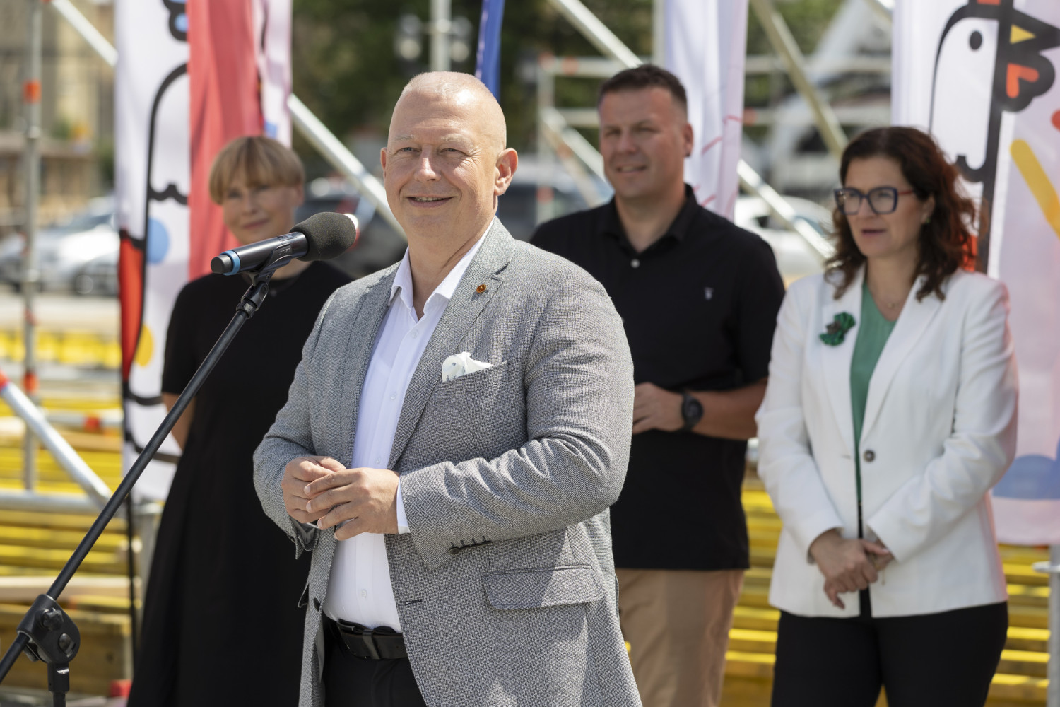Cztery osoby stoją na zewnątrz, ustawione w rzędzie przed mikrofonem. Najbliżej mikrofonu znajduje się mężczyzna w szarym swetrze i białej koszuli, z rękami złożonymi przed sobą. Za nim stoją kolejno: kobieta w czarnej sukience po lewej stronie, mężczyzna w czarnej koszulce polo pośrodku oraz kobieta w białej marynarce i zielonej bluzce po prawej stronie. W tle rozciągają się kolorowe flagi oraz metalowa konstrukcja przypominająca rusztowanie.