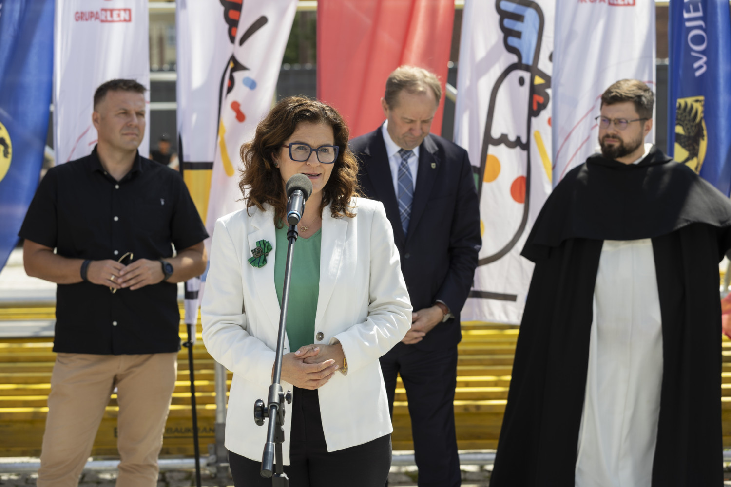 Cztery osoby stoją na zewnątrz przed mikrofonem, otoczone kolorowymi flagami. Na pierwszym planie znajduje się kobieta w białej marynarce i zielonej bluzce, a za nią trzej mężczyźni: jeden w czarnej koszuli, drugi w garniturze, trzeci w stroju duchownego.