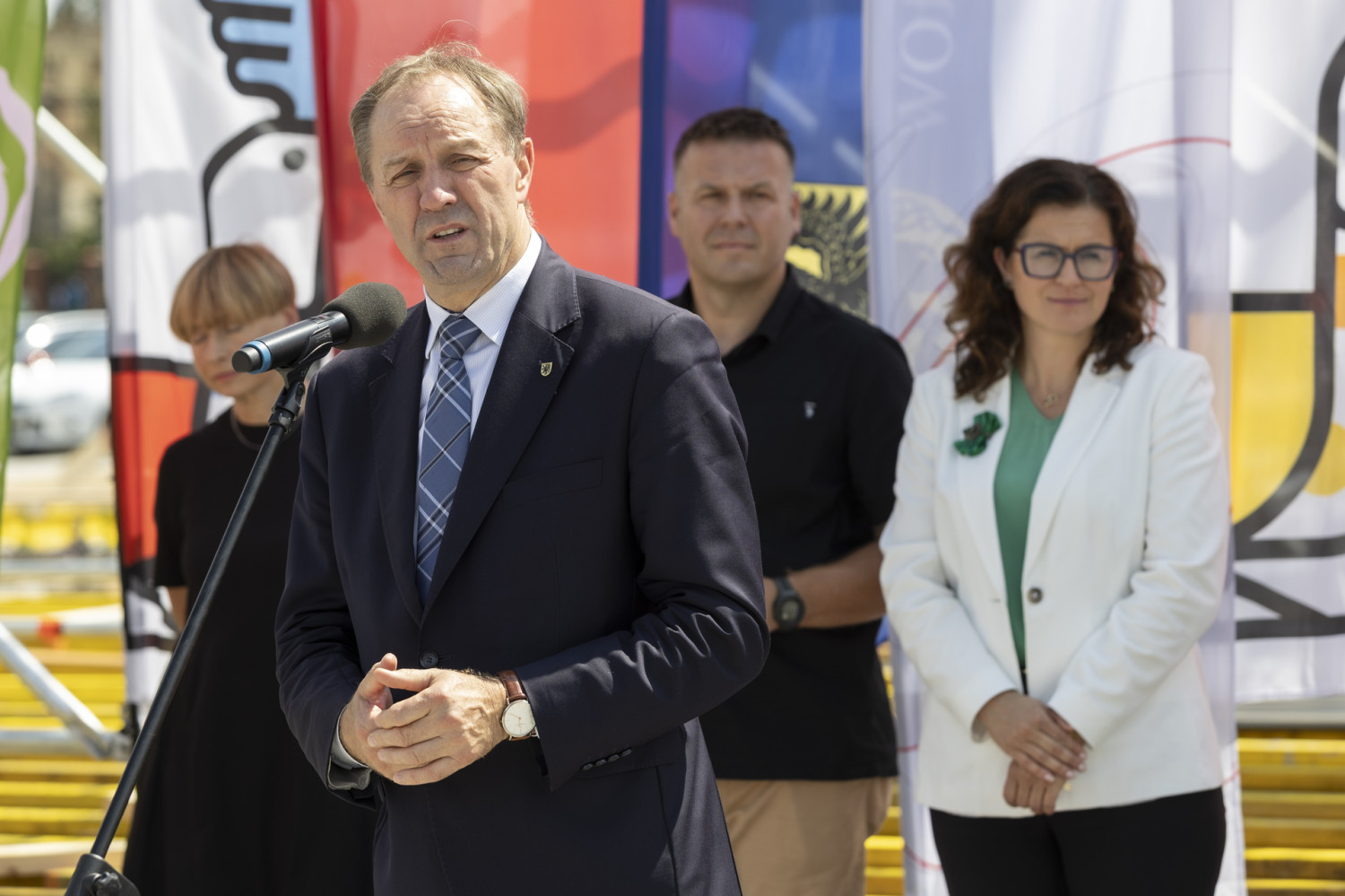 Mężczyzna w garniturze i krawacie przemawia do zgromadzonych, stojąc przy mikrofonie. Za jego plecami ustawione są trzy osoby: kobieta w białej marynarce, mężczyzna w czarnej koszuli oraz kobieta z krótkimi włosami ubrana na czarno. Tło wypełniają kolorowe banery i flagi.